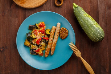 A plate of vegetables with grilled sausages on the table next to zucchini on wooden boards and a wooden fork.