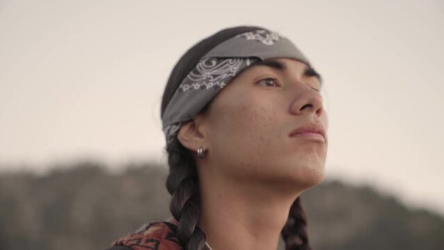 Young Native American man looks off into the distance of a ridge with his chin up. Camera tilts from his face to his hands holding his backpack.