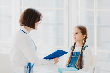 Female pediatrician examines little child, listens carefully to small kid patient, writes down notes in clipboard, pose in clinic or hospital against white window. Children healthcare concept