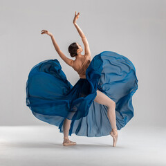 Ballerina in flesh color ballet leotard and blue long ballet skirt dancing in white studio on...