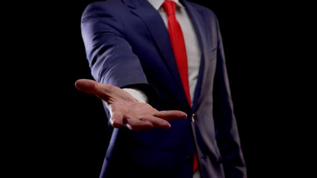 Caucasian businessman in a suit, blue jacket, white shirt with a red tie, stretches hand with open palm helping gesture in the frame closeup, black background, no face visible