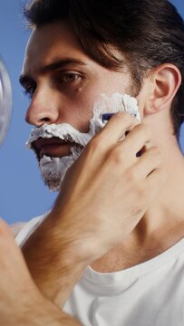 A man shaves his beard looking into a mirror, vertical video on a studio background