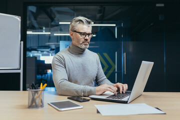 Fototapeta na wymiar Experienced and mature successful businessman focused and serious man working inside office building at laptop, investor in glasses and casual clothes