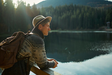 Portrait of young millennial traveller man in hat with backpack enjoying mountain lake with blue...