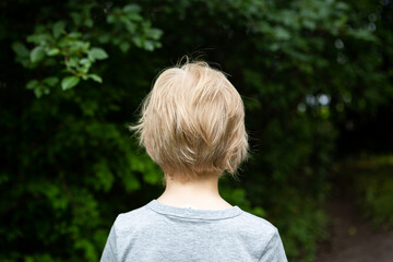 The child's upper body is photographed from behind, with dark green leafy trees in the background....