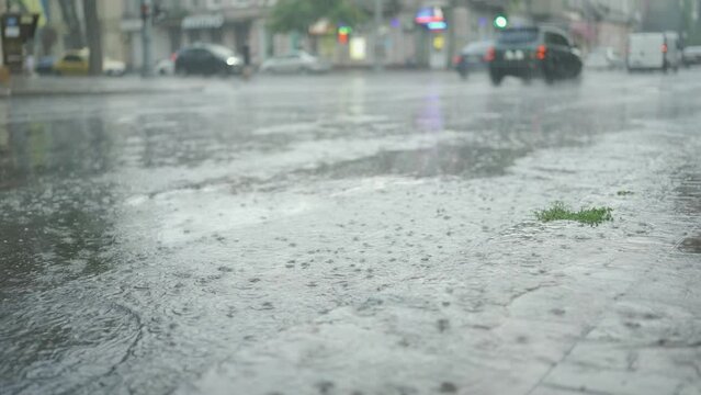 Raindrops fall on the road with cars driving in the background. Heavy torrential rain in the city.