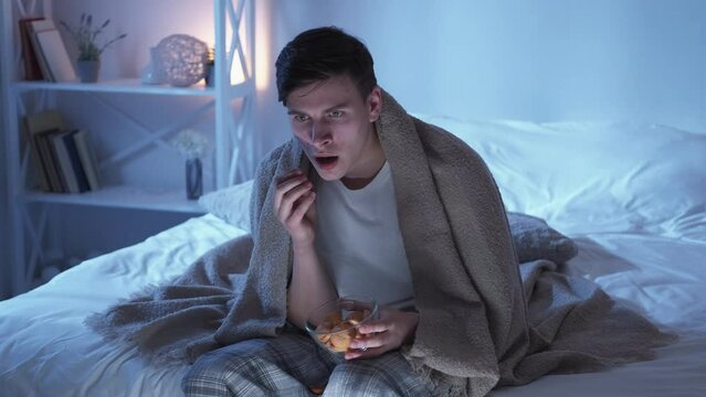 Home Horror Movie. Night Leisure. Weekend Mystery. Shocked Terrified Guy Watching Scary Thriller On TV Eating Snacks In Bed Alone In Bedroom.