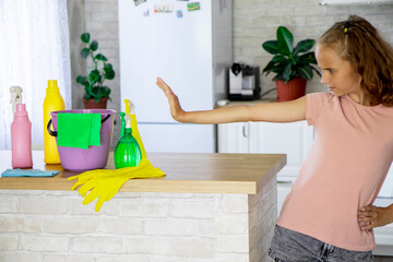 Selective focus on a blonde girl child who shows signs with her hands that she does not want to clean the house. There are detergents, a bucket and rubber gloves on the table.