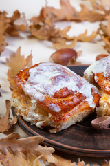 a freshly baked sweet bun on a clay saucer and beautiful autumn oak leaves and acorns in a chaotic order on a white slate or stone background lie in a chaotic order. side view