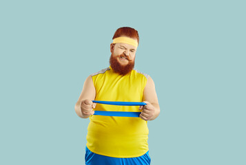 Funny fat redhead man in sportswear isolated on blue studio background exercise train for weight...