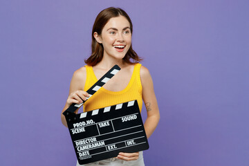 Young woman wear yellow tank shirt hold classic black film making clapperboard look aside on workspace area isolated on plain pastel light purple background studio portrait. People lifestyle concept.