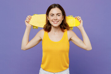 Young smiling happy fun woman 20s she wear yellow tank shirt hold in hand behind neck skateboard pennyboard isolated on plain pastel light purple background studio portrait. People lifestyle concept.