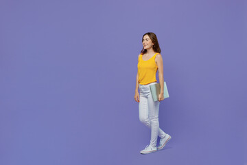 Fototapeta premium Full body side view young funwoman 20s she wear yellow tank shirt hold closed laptop pc computer walking going stroll isolated on plain pastel light purple background studio People lifestyle concept.