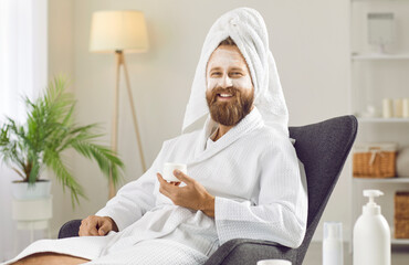 Happy, cheerful, smiling handsome unshaven ginger man in fresh clean white bathrobe and towel enjoying spa and self care day at home and relaxing in comfy chair with kaolin clay spa mask on his face