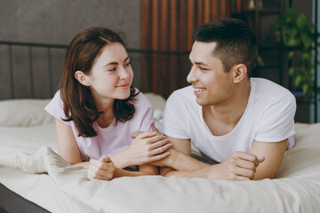 Full body young happy smiling fun couple two family man woman she he wear t-shirt pajama look to each other lying in bed rest spend time together in bedroom home in own room hotel. Real estate concept