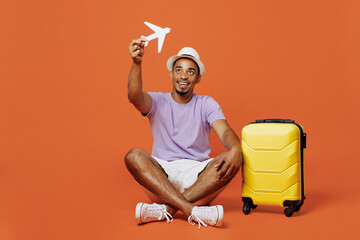 Full body traveler black man wear purple tshirt hat sit hold airplane mockup isolated on plain orange color background Tourist travel abroad in spare time rest getaway Air flight trip journey concept