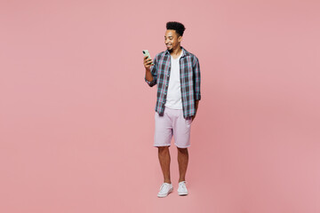 Fototapeta premium Full body young smiling man of African American ethnicity he wear blue shirt hold in hand use mobile cell phone isolated on plain pastel light pink background studio portrait People lifestyle concept