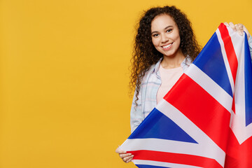 Fototapeta premium Young smiling happy fun black teen girl student she wears casual clothes backpack bag hold british flag look camera isolated on plain yellow color background. High school university college concept.