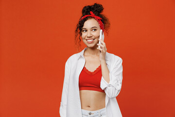 Young happy woman of African American ethnicity she wear white shirt top talk speak on mobile cell phone conducting pleasant conversation isolated on plain orange background. People lifestyle concept.