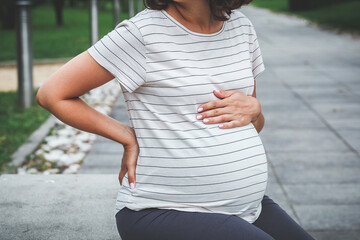 a pregnant woman sits in a park on a belly, holding her stomach and back, discomfort and pain...