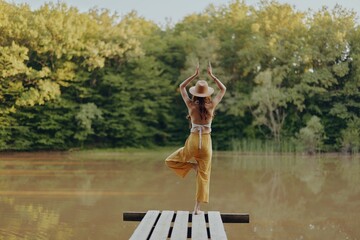 A woman stands on a bridge on a lake and meditates holding her balance on one leg in a hippie image...