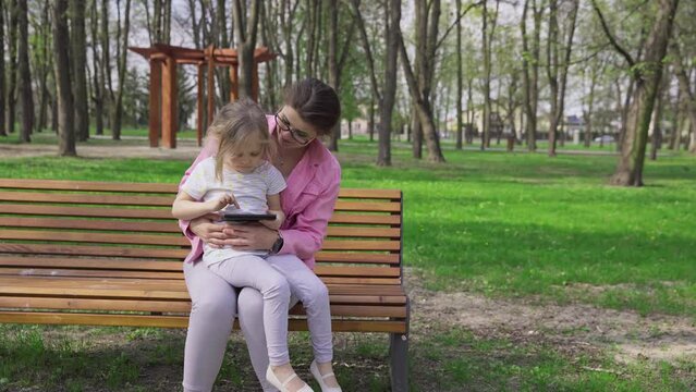 A mother and child sit on a bench and both look at a smartphone.