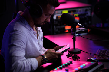 Young man host enjoying podcasting in studio, speaking into a microphone, holding a phone, using laptop. Handsome podcaster streaming live audio podcast