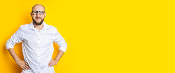 Smiling young bearded man in glasses holding hands on waist on yellow backgroun. Banner