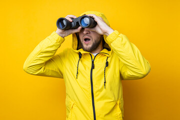 Surprised young man in yellow raincoat looking through binoculars on yellow background