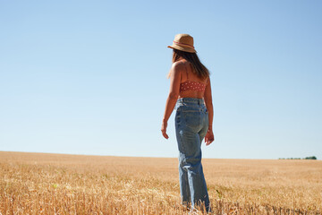Naklejka na ściany i meble A beautiful young woman standing in a wheat field on a sunny day in a countryside