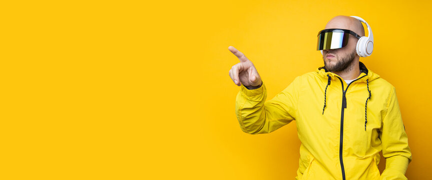 Young man in cyberpunk glasses with headphones clicking on an invisible screen on a yellow background. Banner