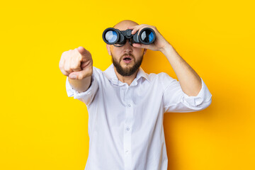 Surprised young man looking through binoculars pointing his finger in front on yellow background