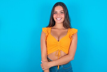 brunette woman wearing orange tank top over blue background  with broad smile, shows white teeth,...