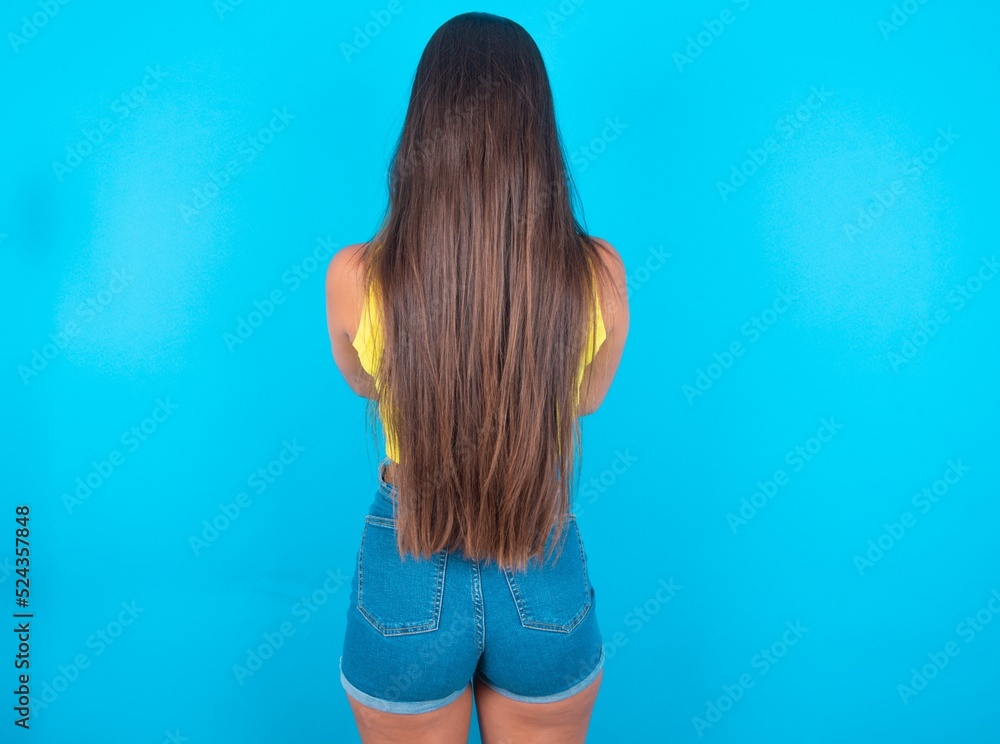 Wall mural brunette woman wearing yellow tank top over blue background standing backwards looking away with arms on body.