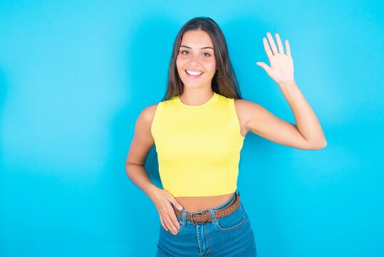 brunette woman wearing yellow tank top over blue background waiving saying hello or goodbye happy and smiling, friendly welcome gesture.