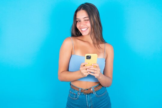 Pleased beautiful brunette woman wearing blue tank top over blue background using self phone and looking and winking at the camera. Flirt and coquettish concept.