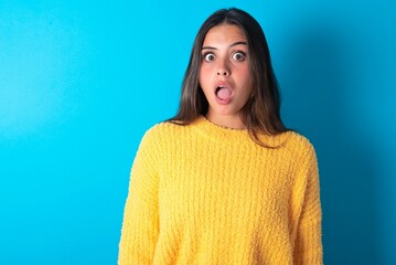 Shocked beautiful brunette woman wearing yellow sweater over blue background stares bugged eyes...