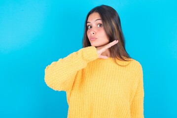 beautiful brunette woman wearing yellow sweater over blue background cutting throat with hand as...