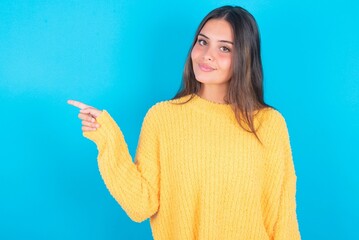 beautiful brunette woman wearing yellow sweater over blue background points to side on blank space demonstrates advertisement. People and promotion concept