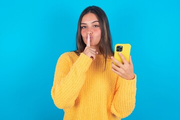 beautiful brunette woman wearing yellow sweater over blue background holding modern gadget ask not tell secrets