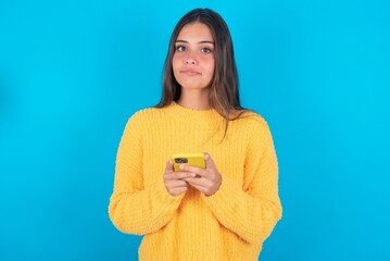 Portrait of serious confident beautiful brunette woman wearing yellow sweater over blue background...