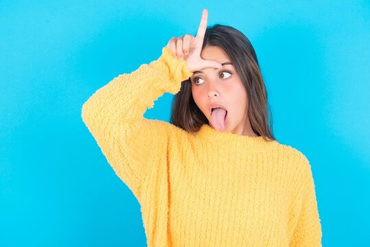 Funny beautiful brunette woman wearing yellow sweater over blue background makes loser gesture mocking at someone sticks out tongue making grimace face.