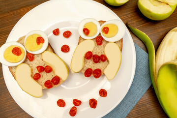 Funny owl face sandwich toast bread with chicken eggs, apple, banana, dried berry fruits on plate. Cute kids childrens baby's sweet dessert breakfast,lunch, food art on wooden background,top view