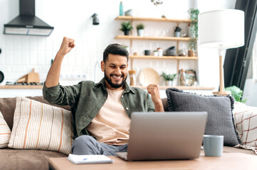 Amazed excited arabian or indian man, freelancer, sits on a sofa in living room, looks at laptop...