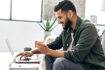 Side view of satisfied positive indian or arabian guy in stylish clothes sitting on a sofa in a living room, using laptop and bank card to pay online, ordering goods and clothes, home delivery, smiles