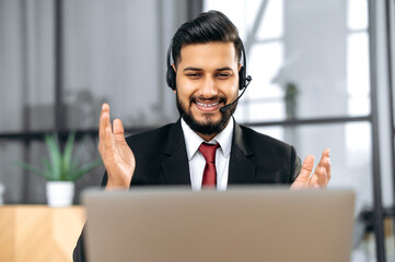 Online video negotiations. Positive friendly indian or arabian young business man, mentor, coach, with headset, looking at laptop screen, gesturing with hands, talking to colleagues or students, smile