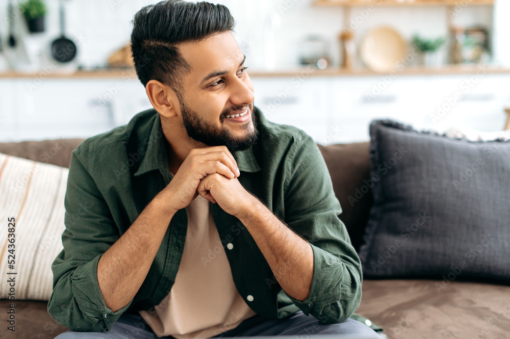 Wall mural Handsome positive confident attractive indian or arabian man, in casual stylish clothes, sitting at home in cozy living room on the background of the kitchen, looks to the side, smiling