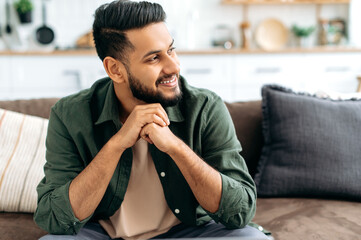 Handsome positive confident attractive indian or arabian man, in casual stylish clothes, sitting at home in cozy living room on the background of the kitchen, looks to the side, smiling
