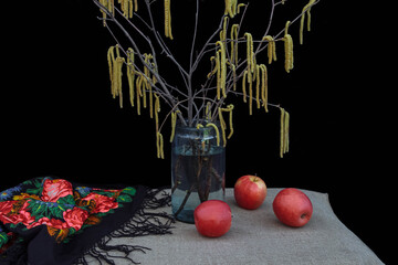 Retro still life with vintage shawl and a bouquet of spring birch branches with buds  in vase and red apples on  table  black background, concept of old rustic interiors, winter, arrival of Carnival