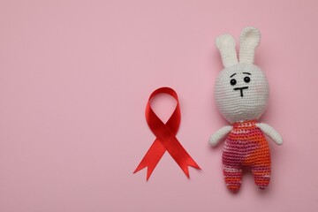 Cute knitted toy bunny and red ribbon on pink background, flat lay with space for text. AIDS disease awareness